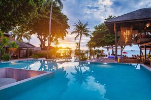 A resort pool with clear water, surrounded by palm trees and lounge chairs, with a view of the sunset over the ocean.