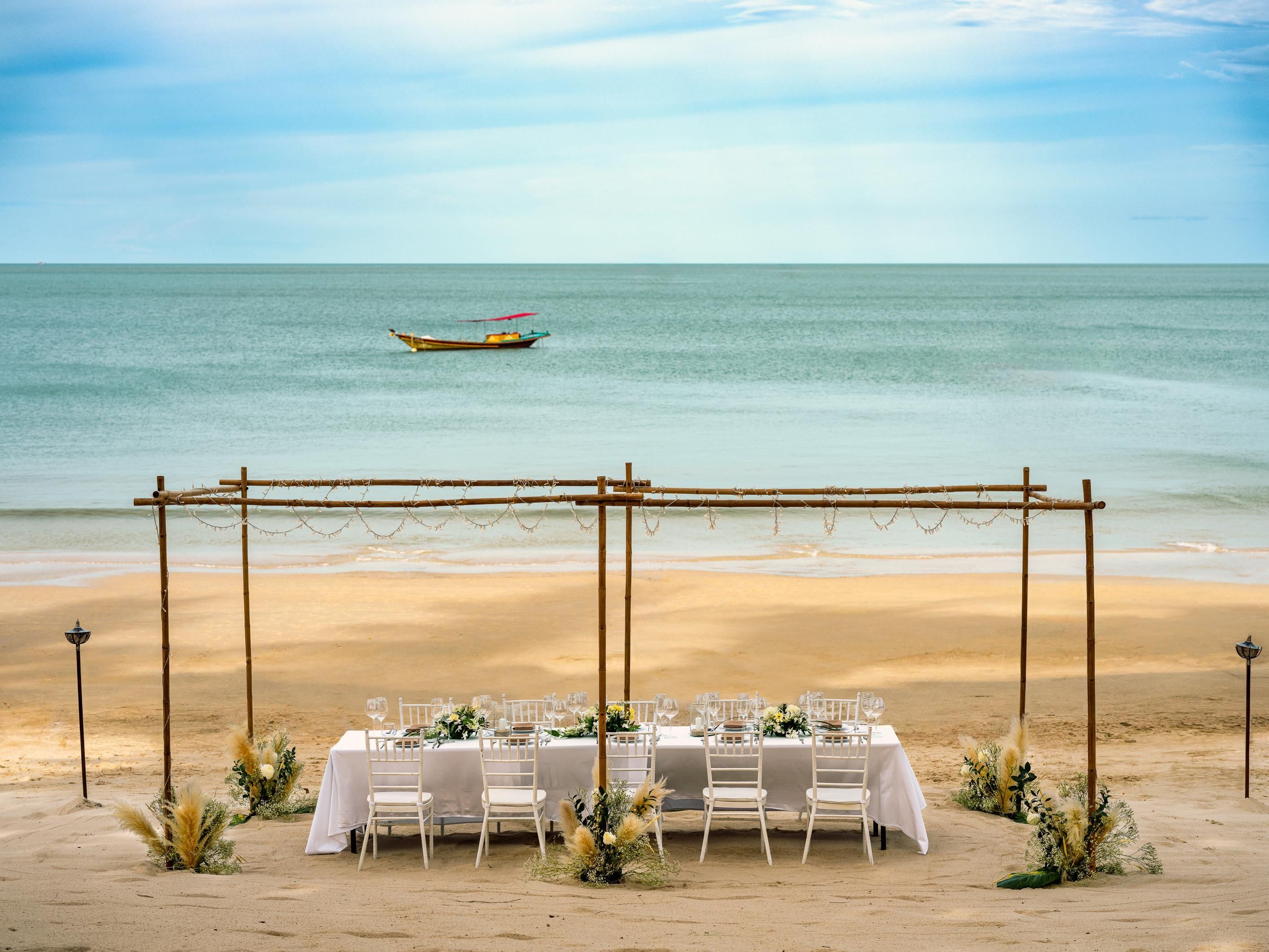A beachside dining setup with a table and chairs, a wooden pergola, and a boat in the background.