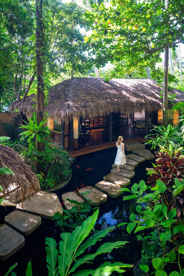 A tropical resort with thatched-roof structures, a water feature, and a person standing on a stone bridge.