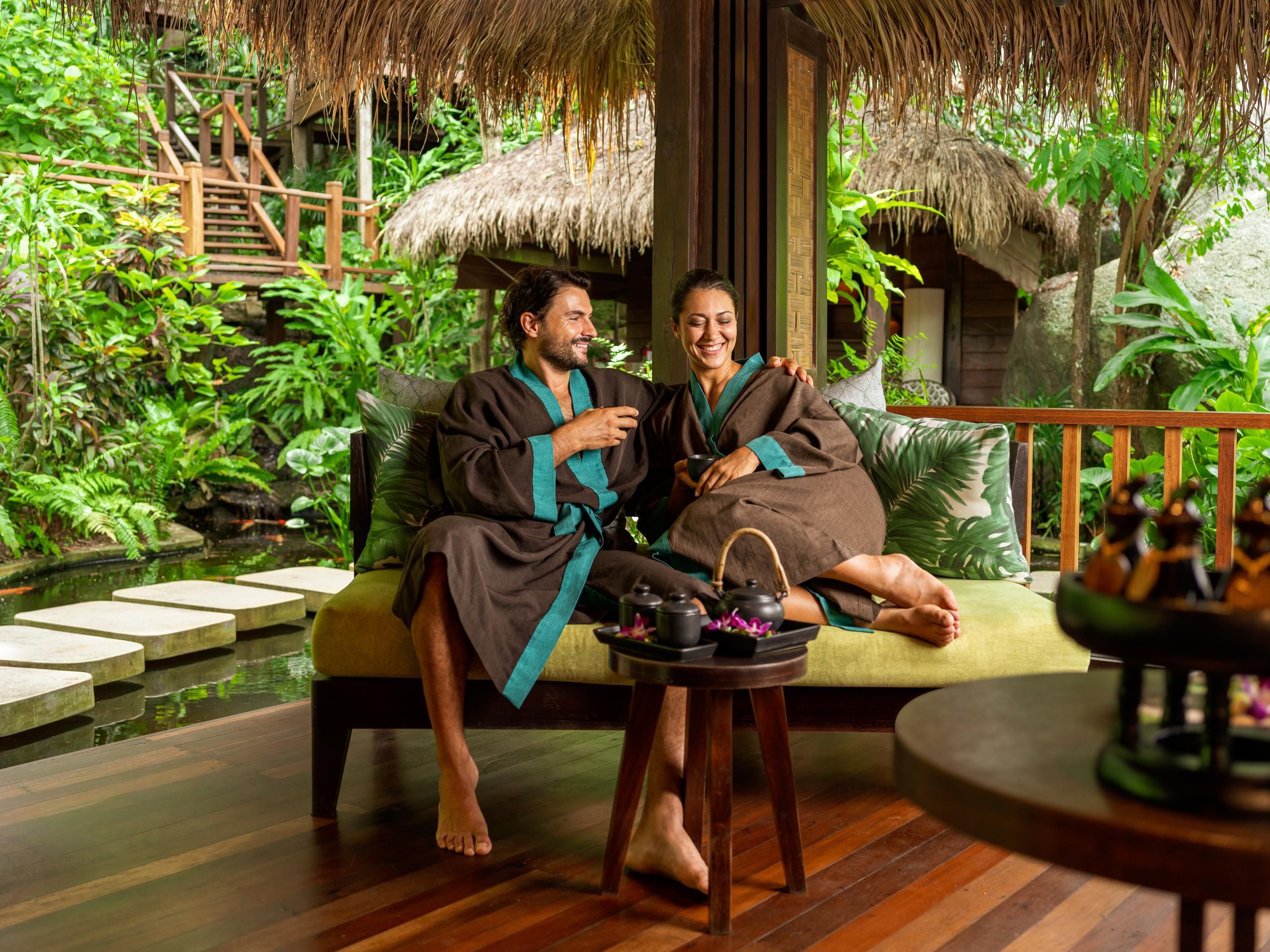 A couple seated on a wooden bench with a thatched roof, surrounded by lush greenery and a small pond.