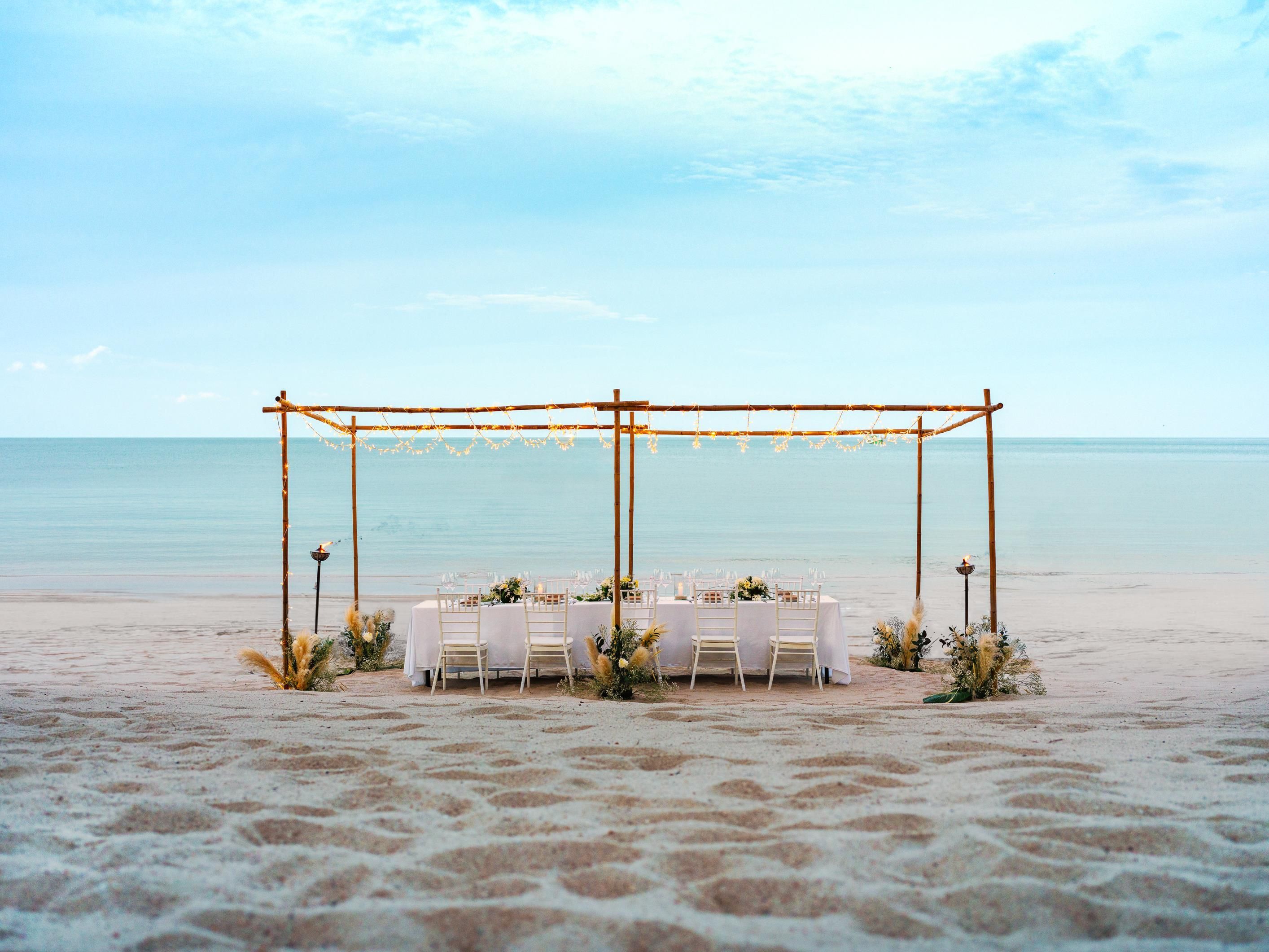 A beach wedding setup with a wooden arch, tables, chairs, and string lights.