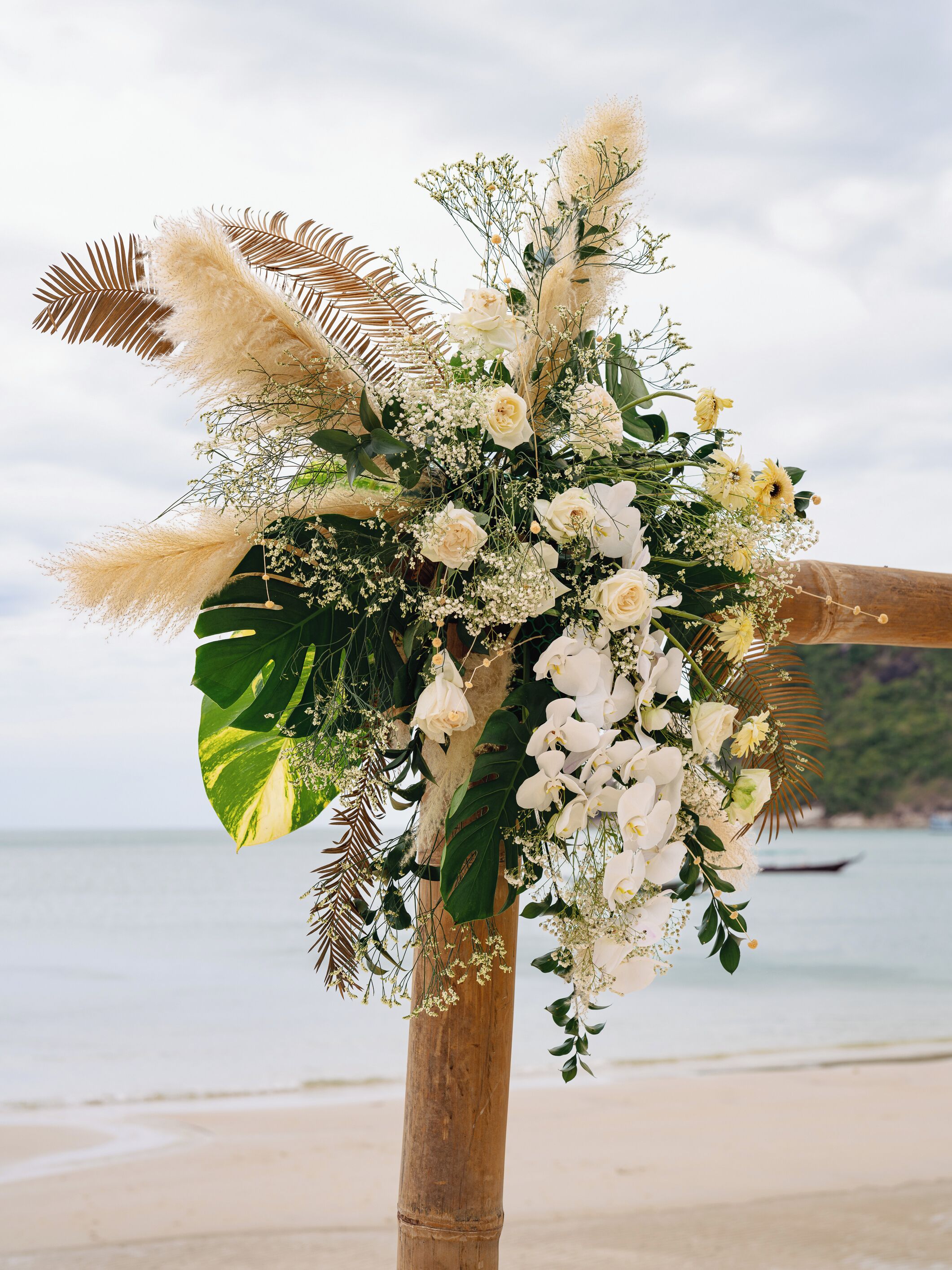 Um arco de casamento decorado com flores brancas e capim-do-campo, com o mar ao fundo.