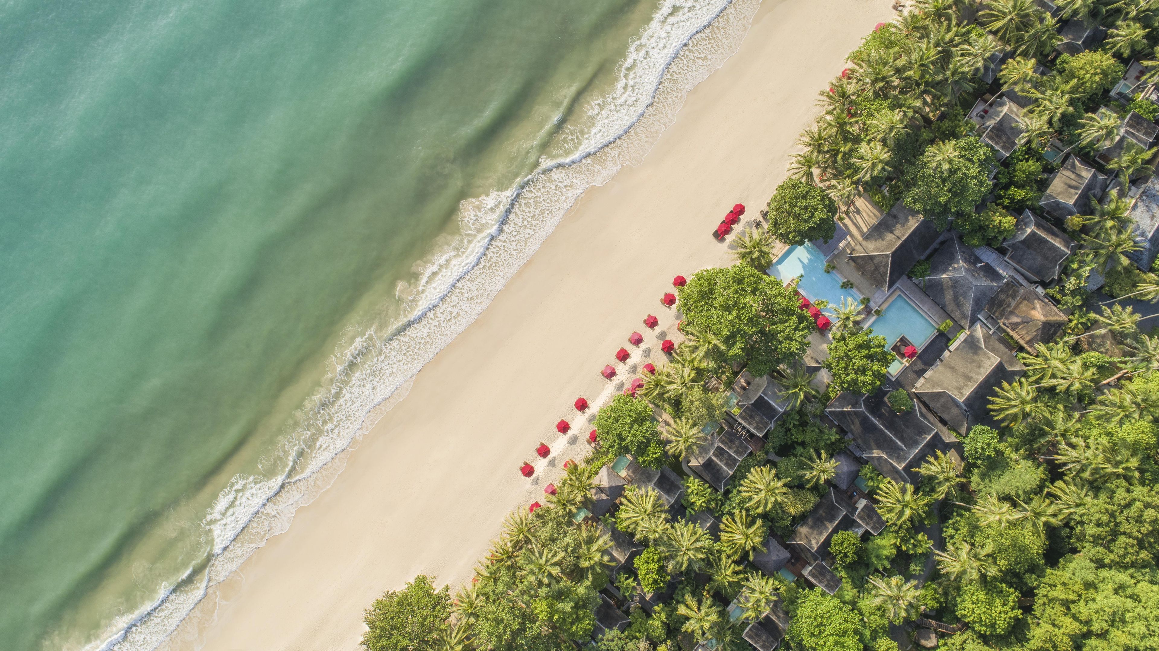 An aerial view of a beach resort with a swimming pool, red umbrellas, and palm trees.