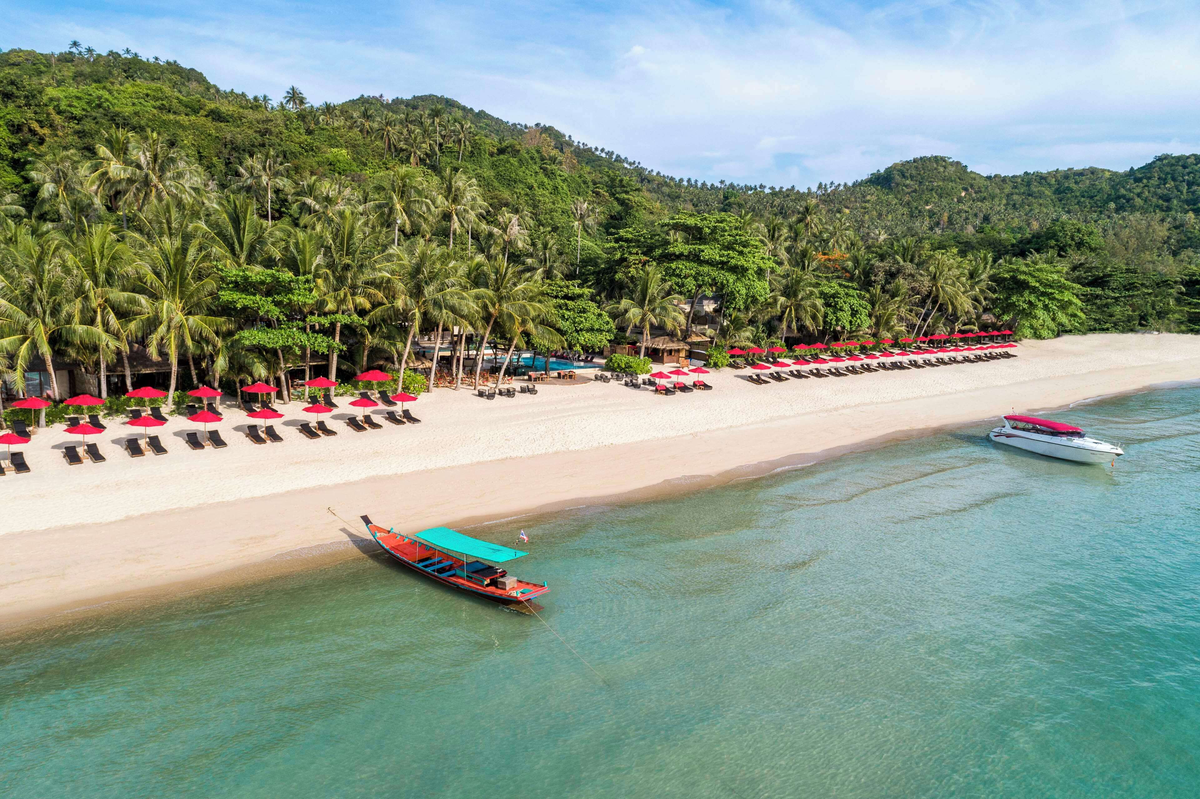 A beach with rows of sun loungers and umbrellas, a boat, and a forested hill in the background.