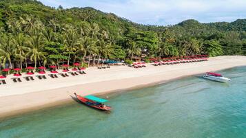 A beach with rows of sun loungers and umbrellas, a boat, and a forested hill in the background.