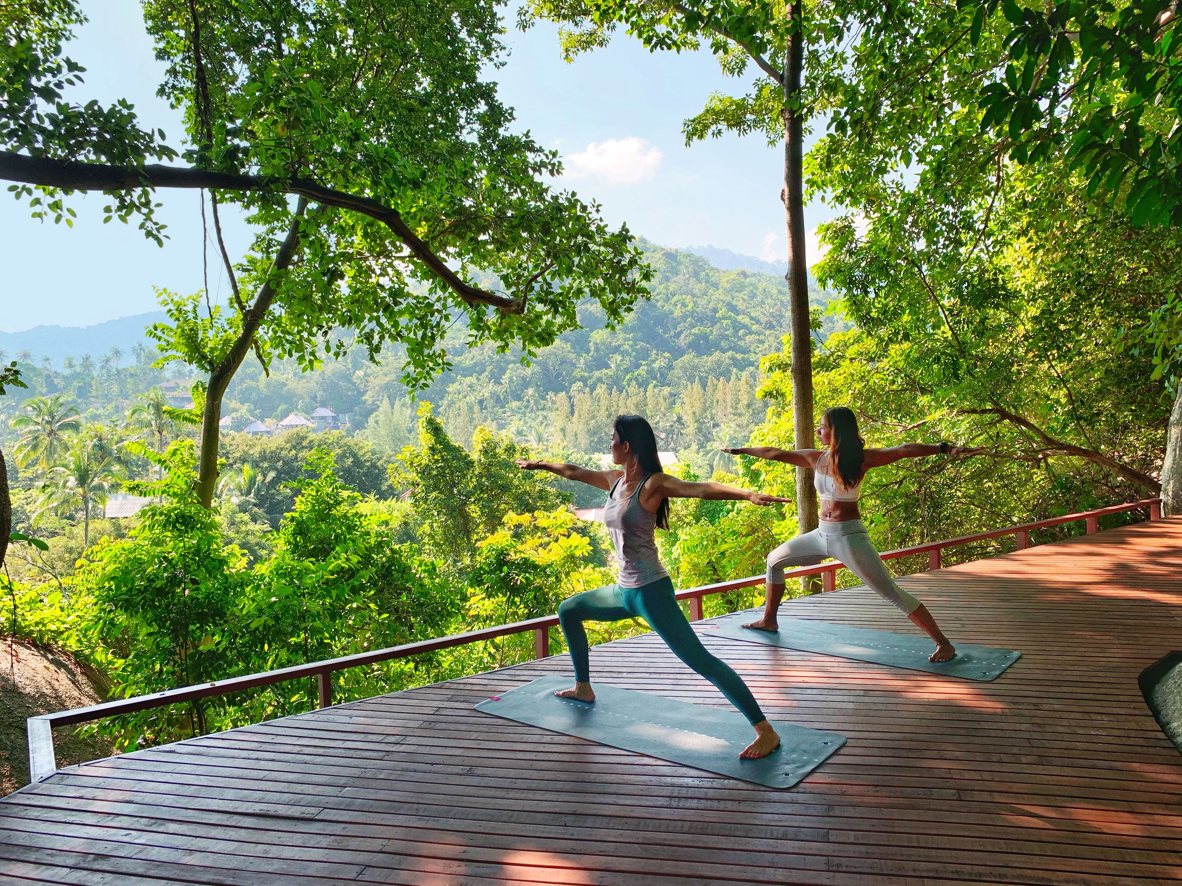 Duas pessoas praticando ioga num deck de madeira, com uma vista panorâmica de uma paisagem verdejante e montanhas ao fundo.