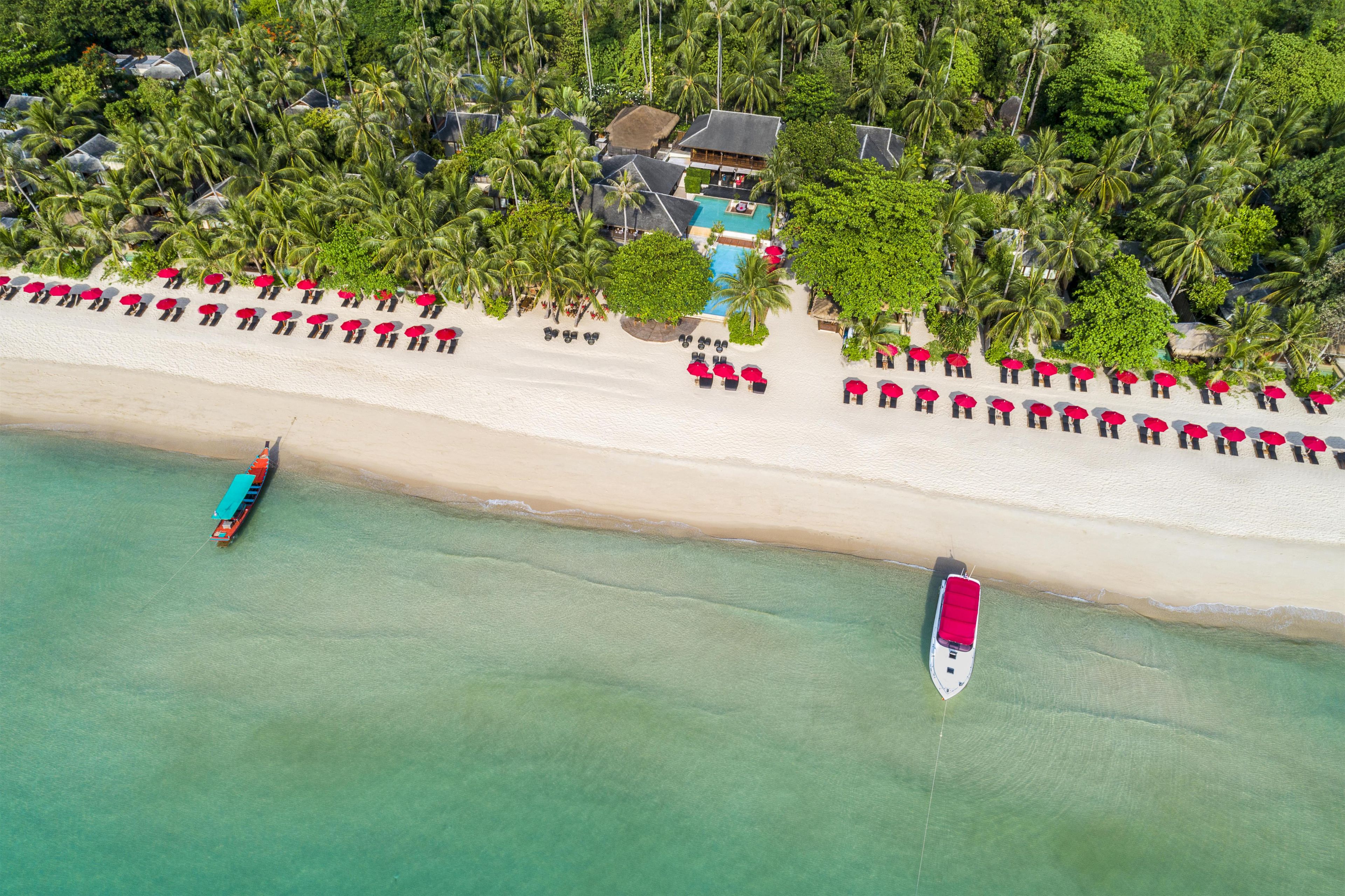 An aerial view of a beach resort with a swimming pool, lounge chairs, and boats.