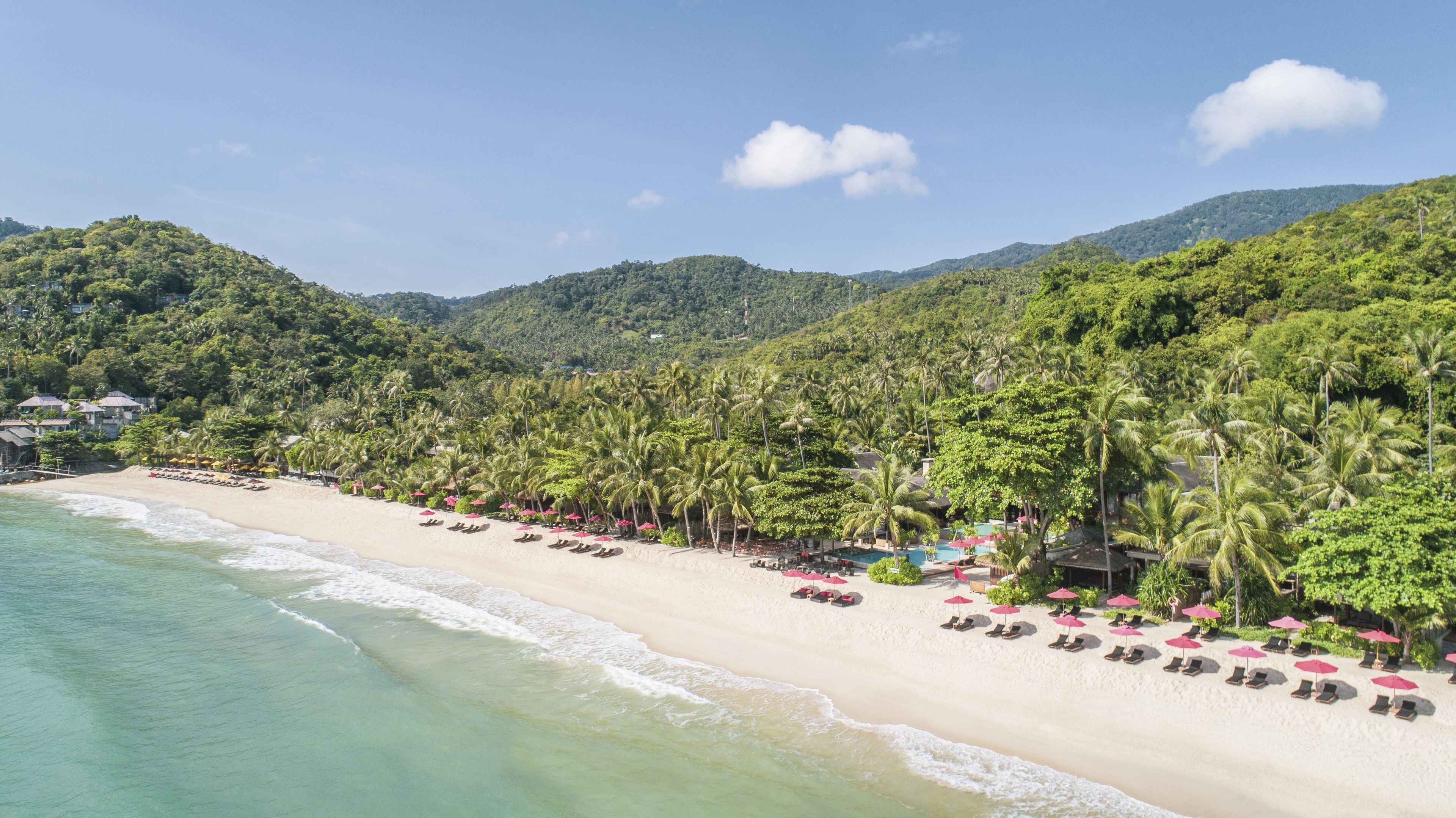 A beach with rows of sun loungers and umbrellas, a pool, and a backdrop of lush green hills.