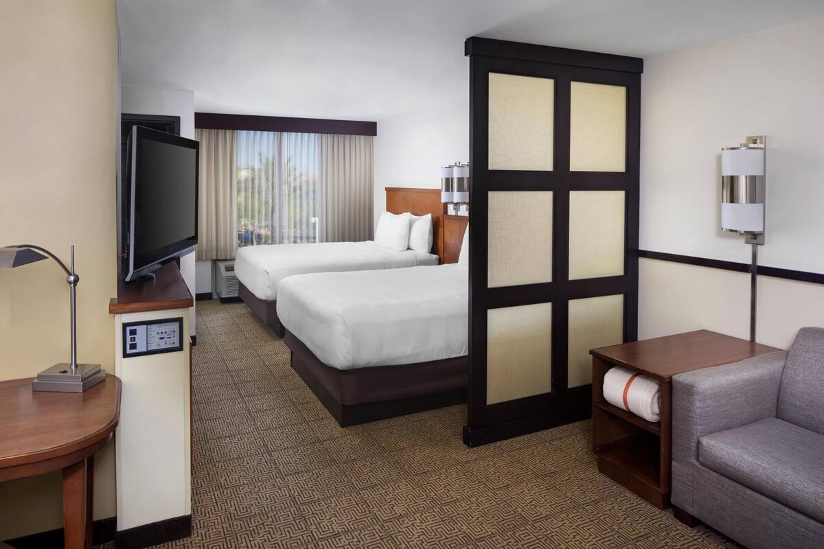 A hotel room with two beds, a TV, a desk, a chair, and a large window with curtains.