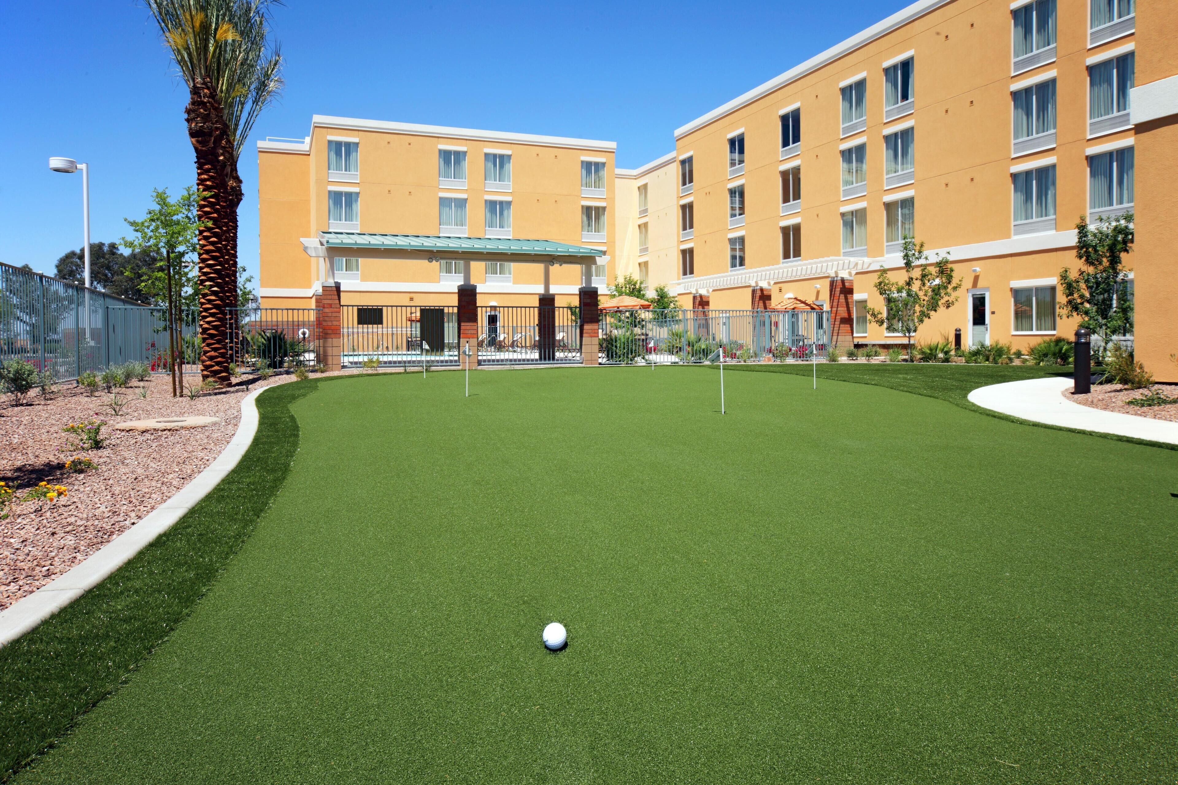 A golf course with a putting green, a palm tree, and a building with multiple windows and balconies in the background.