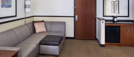 A hotel room with a gray sofa, a small table with a towel, a patterned carpet, a wooden door, a kitchenette with a coffee maker, and a framed picture on the wall.
