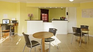 A compact kitchen with a dining area, featuring a round wooden table, two chairs, a small island with a sink, and a computer on a desk. - Americas Best Value Inn Pryor (Pryor)