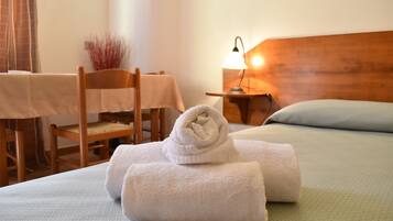 Una cama bien hecha con dos toallas enrolladas, una silla de madera, una mesita con mantel y una lámpara de noche.