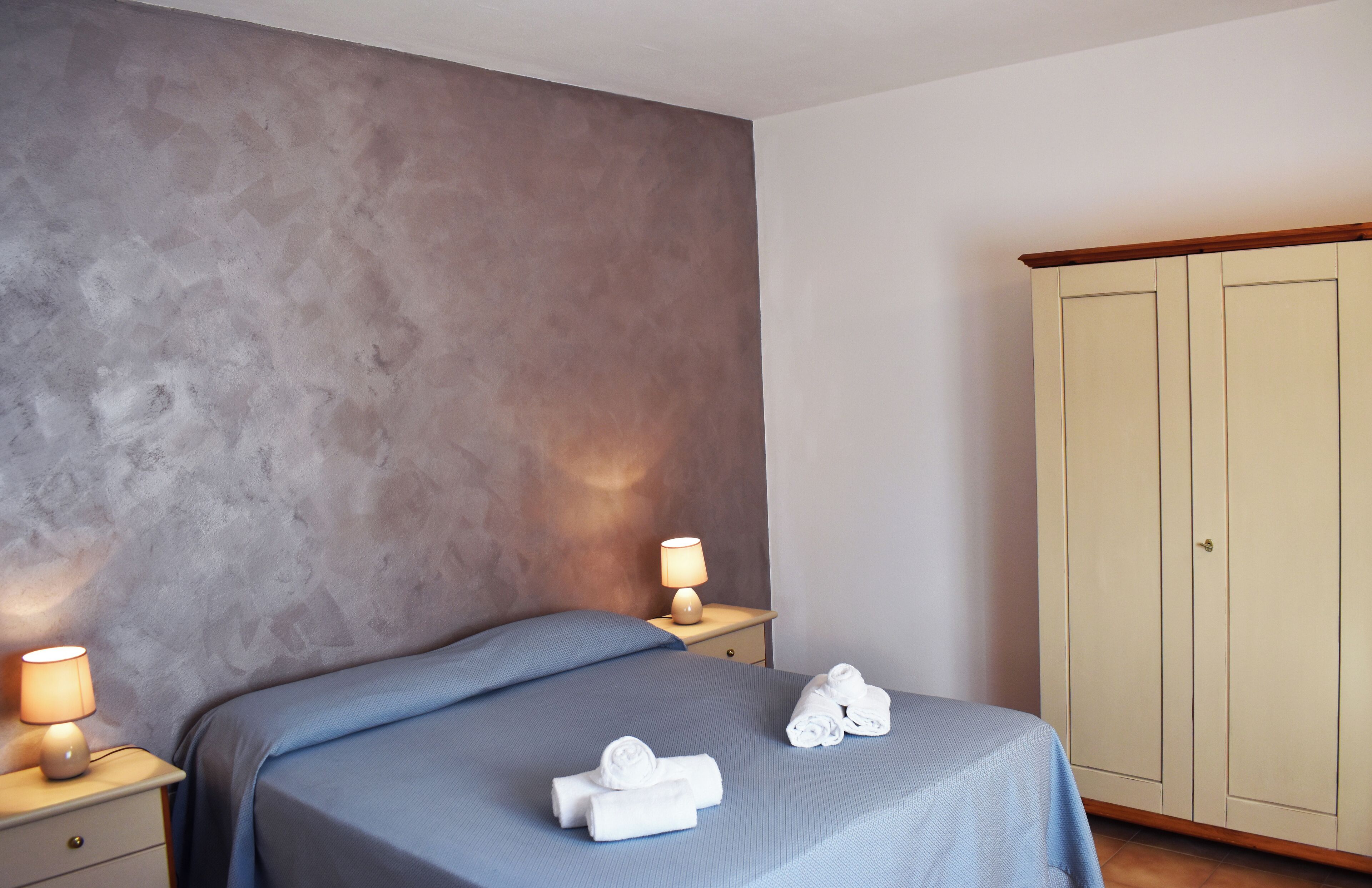 Un dormitorio con cama, mesita de noche con lámpara, armario y dos toallas sobre la cama.