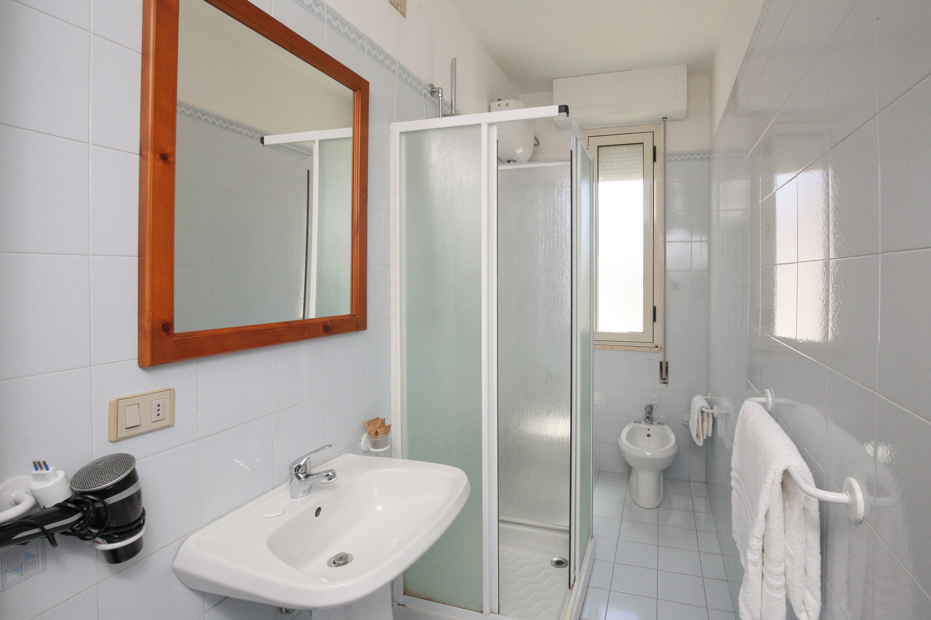 Un baño con un lavamanos blanco, un espejo, cabina de ducha de vidrio, un bidé y un perchero para toallas.