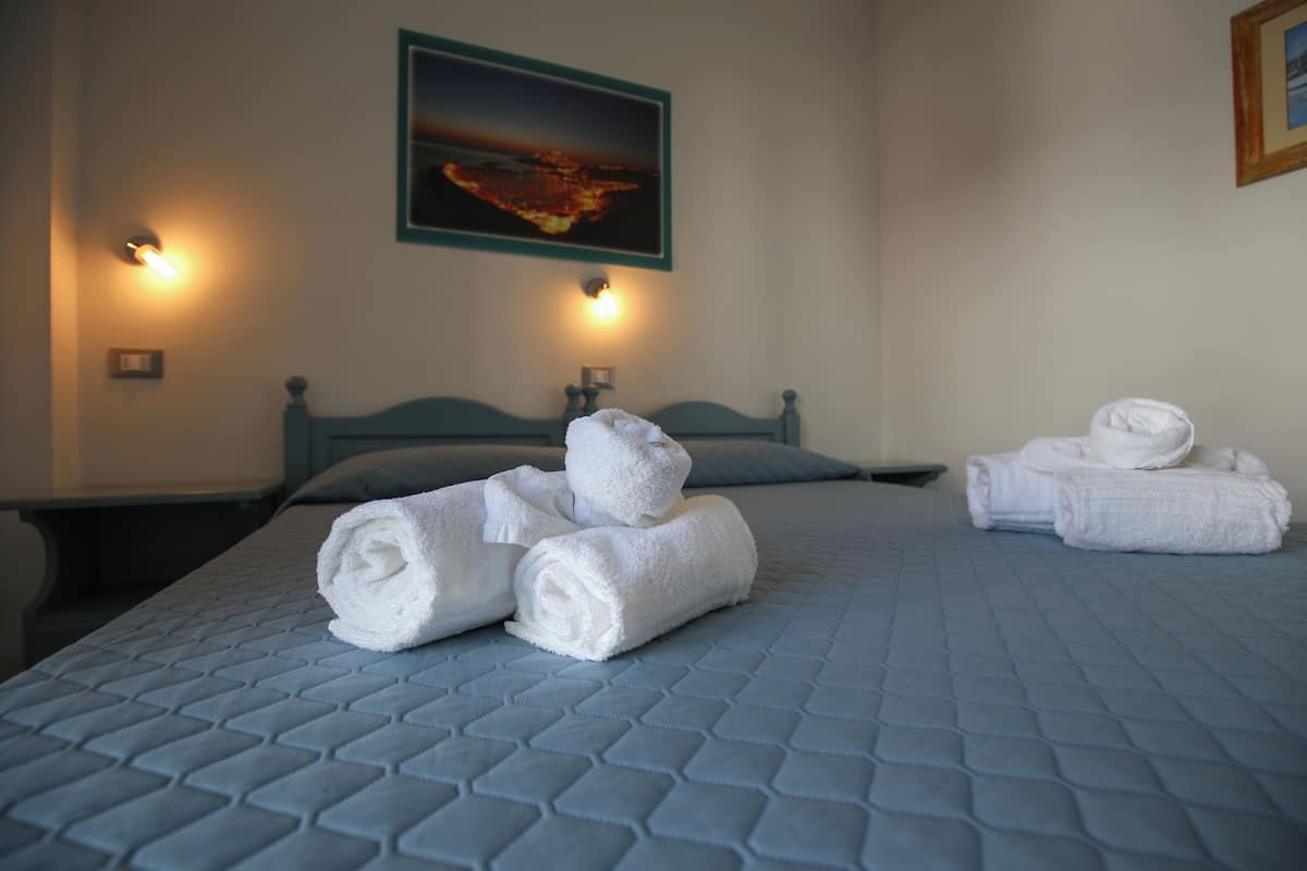 A neatly made bed with rolled white towels, a cityscape painting on the wall, and two wall-mounted light fixtures.