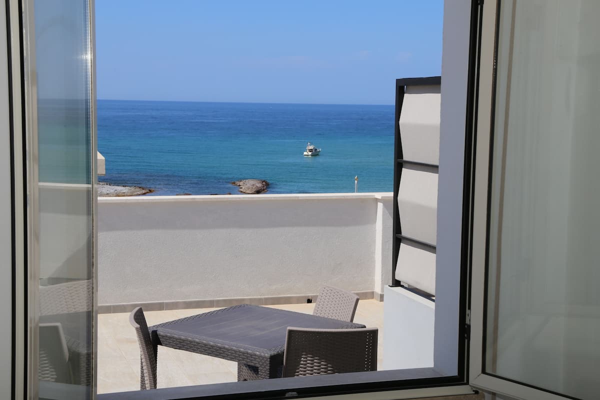 A balcony with a table and chairs, overlooking a calm sea with a boat and rocks.