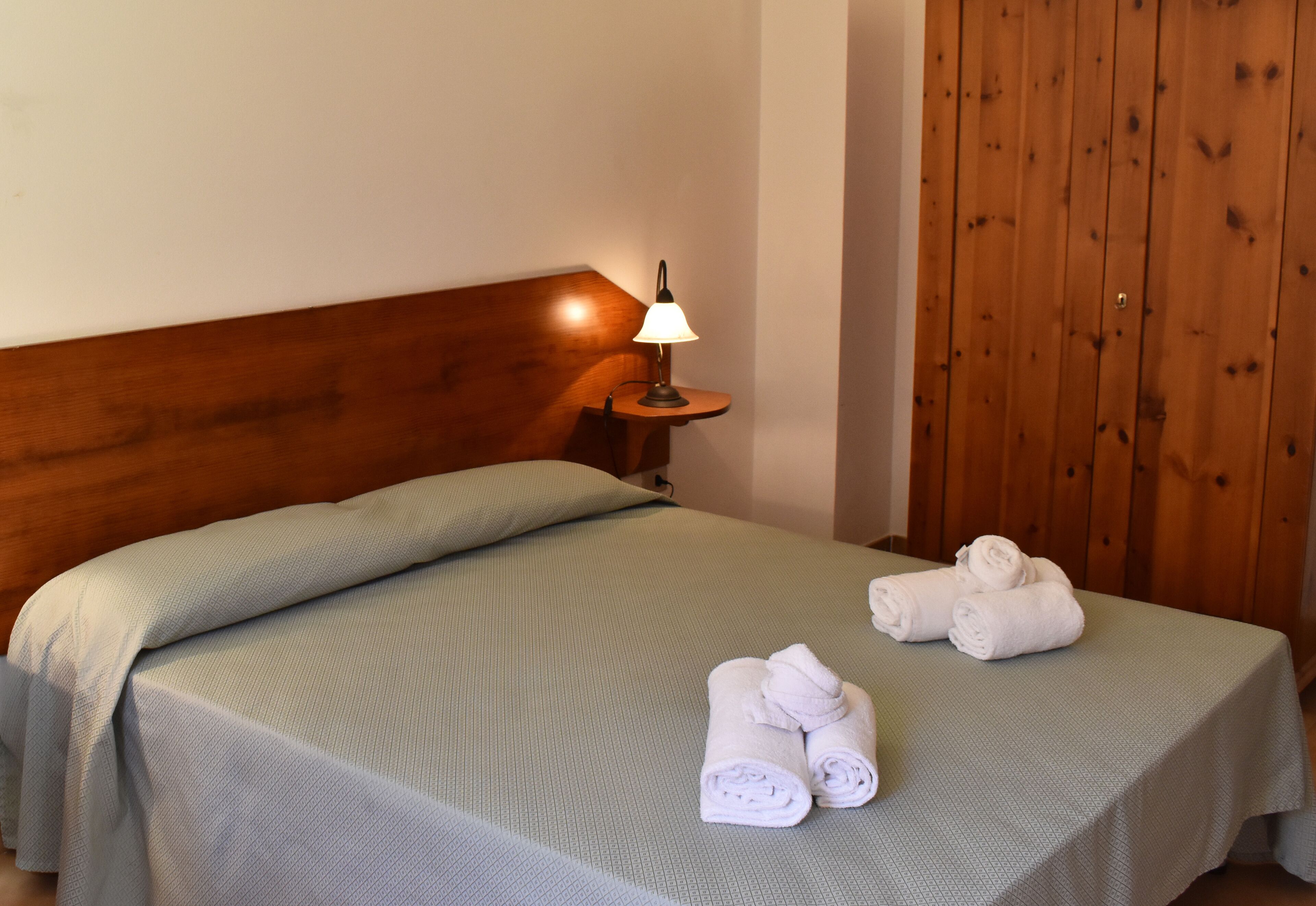 A neatly made bed with a green bedspread, a bedside lamp, and two rolled towels on the bed.