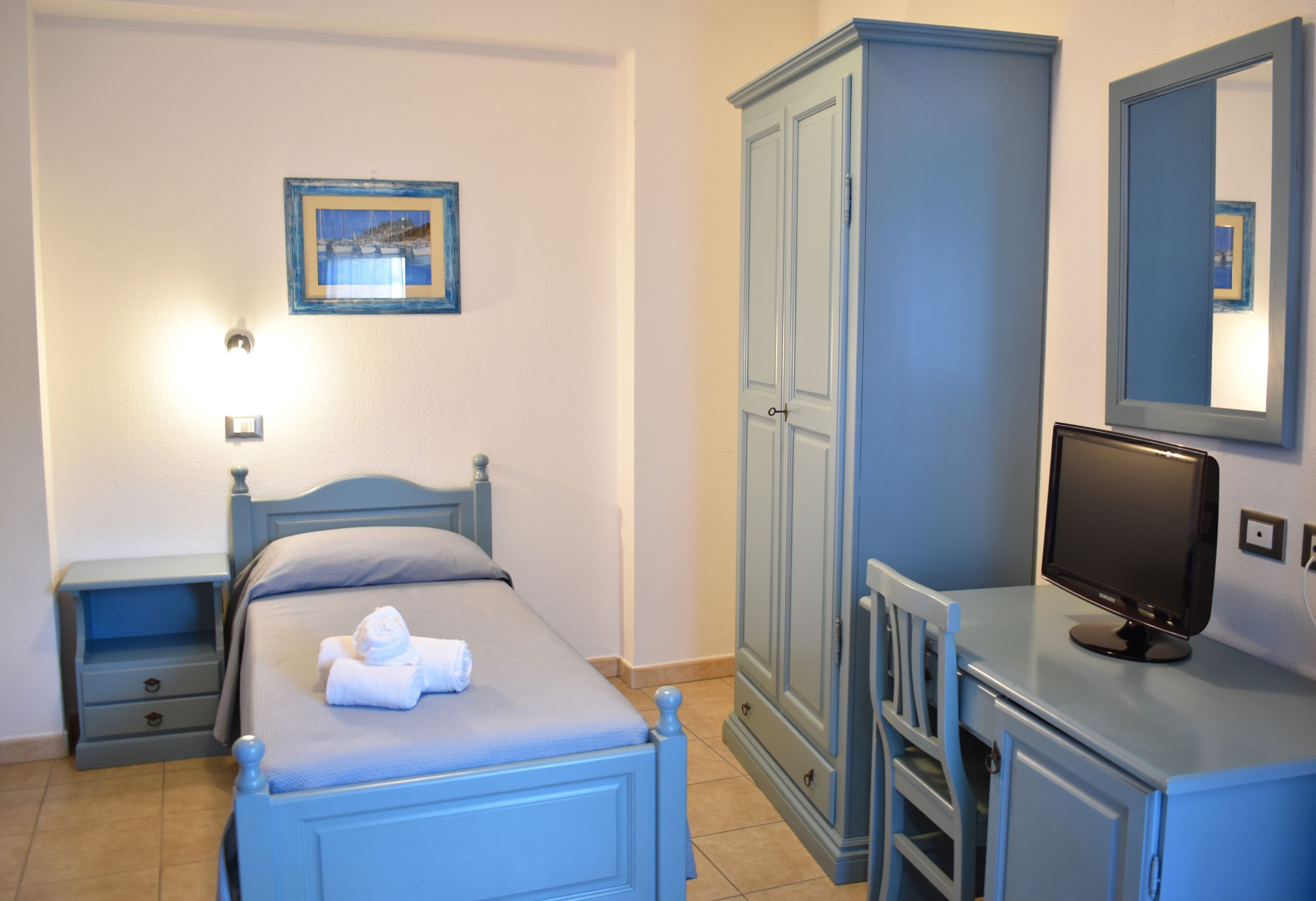 A bedroom with a bed, a nightstand, a wardrobe, a desk with a computer, and a mirror.