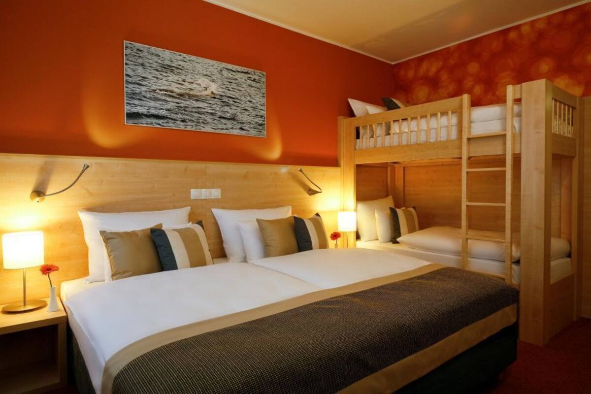 Habitación de hotel con una cama grande, dos mesitas de noche con lámparas, una litera de madera y un cuadro en la pared.