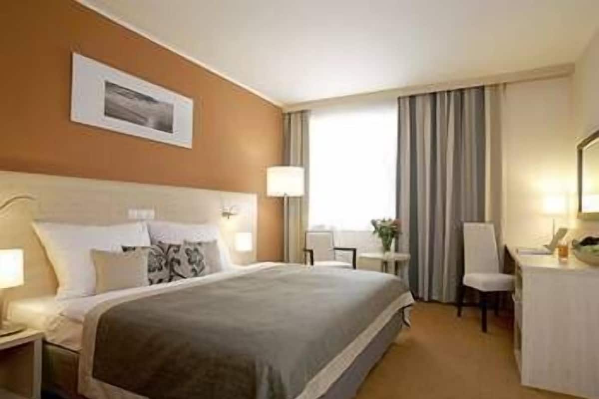 Habitación de hotel con una cama grande, un escritorio, una silla y un ventanal con cortinas.