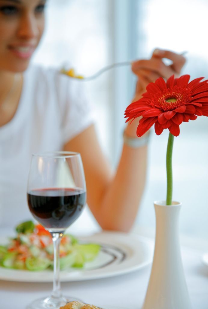 A dining table with a glass of red wine, a plate of food, and a vase with a red flower.