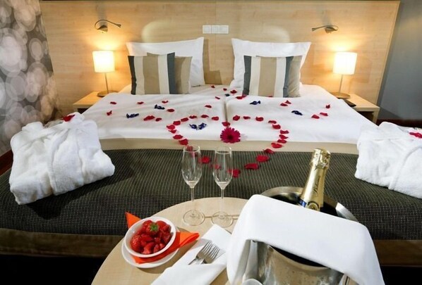 A hotel room with a bed, two bedside lamps, a bedside table with a bottle of champagne, a bowl of strawberries, and a plate of strawberries. - Aquapalace Hotel Prague (Cestlice)