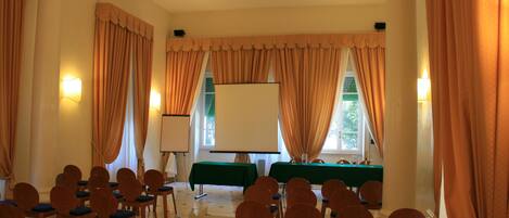 Uma sala de conferências com fileiras de cadeiras de madeira, uma tela de projetor e uma mesa com toalha verde.