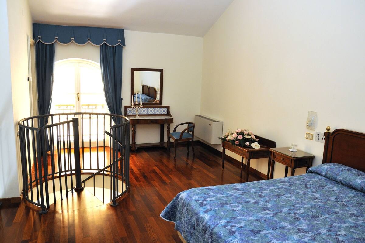 Une chambre avec un plancher en bois, un lit recouvert d’une couvre-lit à motifs floraux, un bureau avec une chaise, une petite table ornée de fleurs et une fenêtre avec des rideaux.
