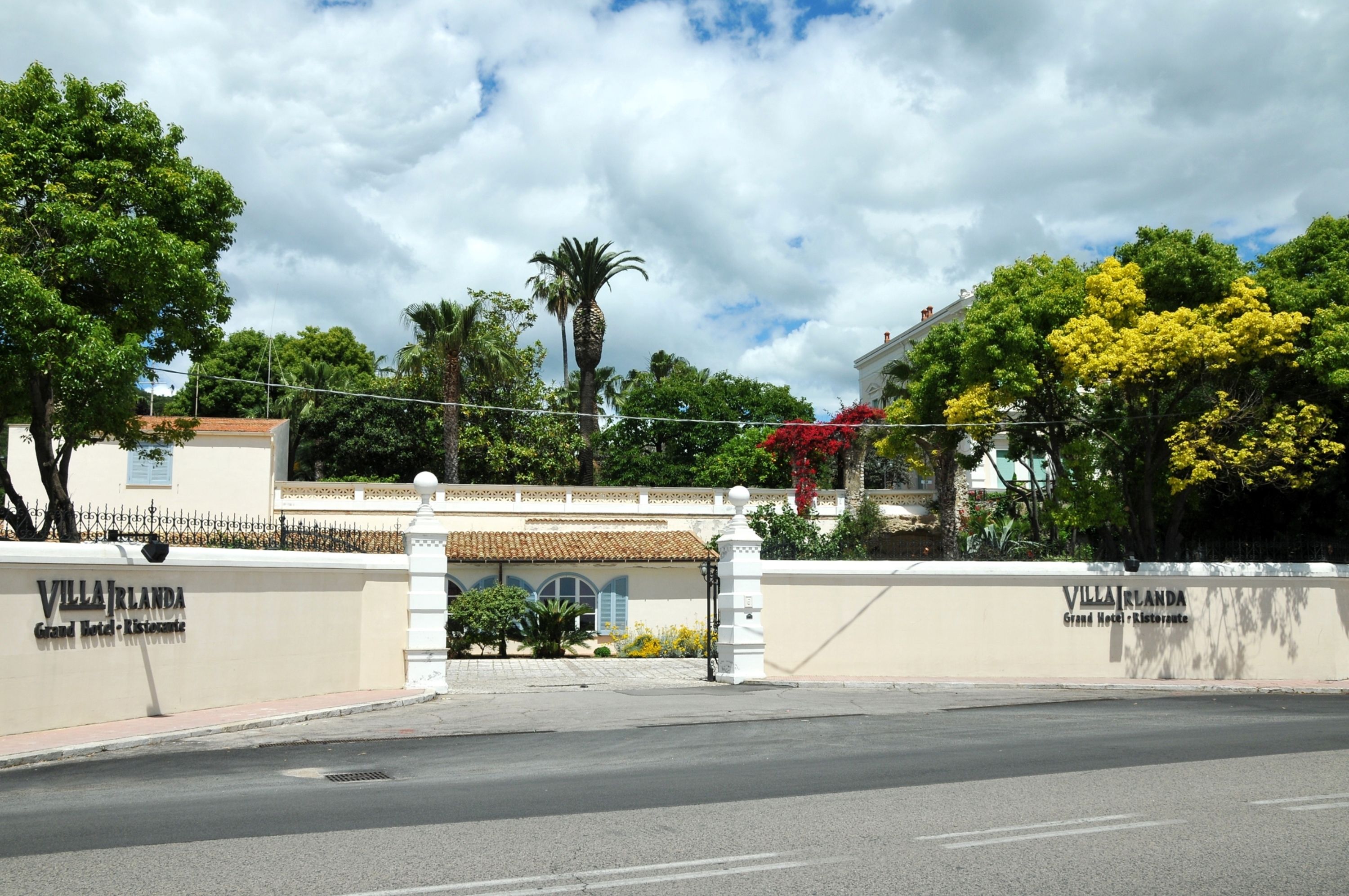Villa Irlanda's entrance with a white wall, a palm tree, and a red flowering plant.