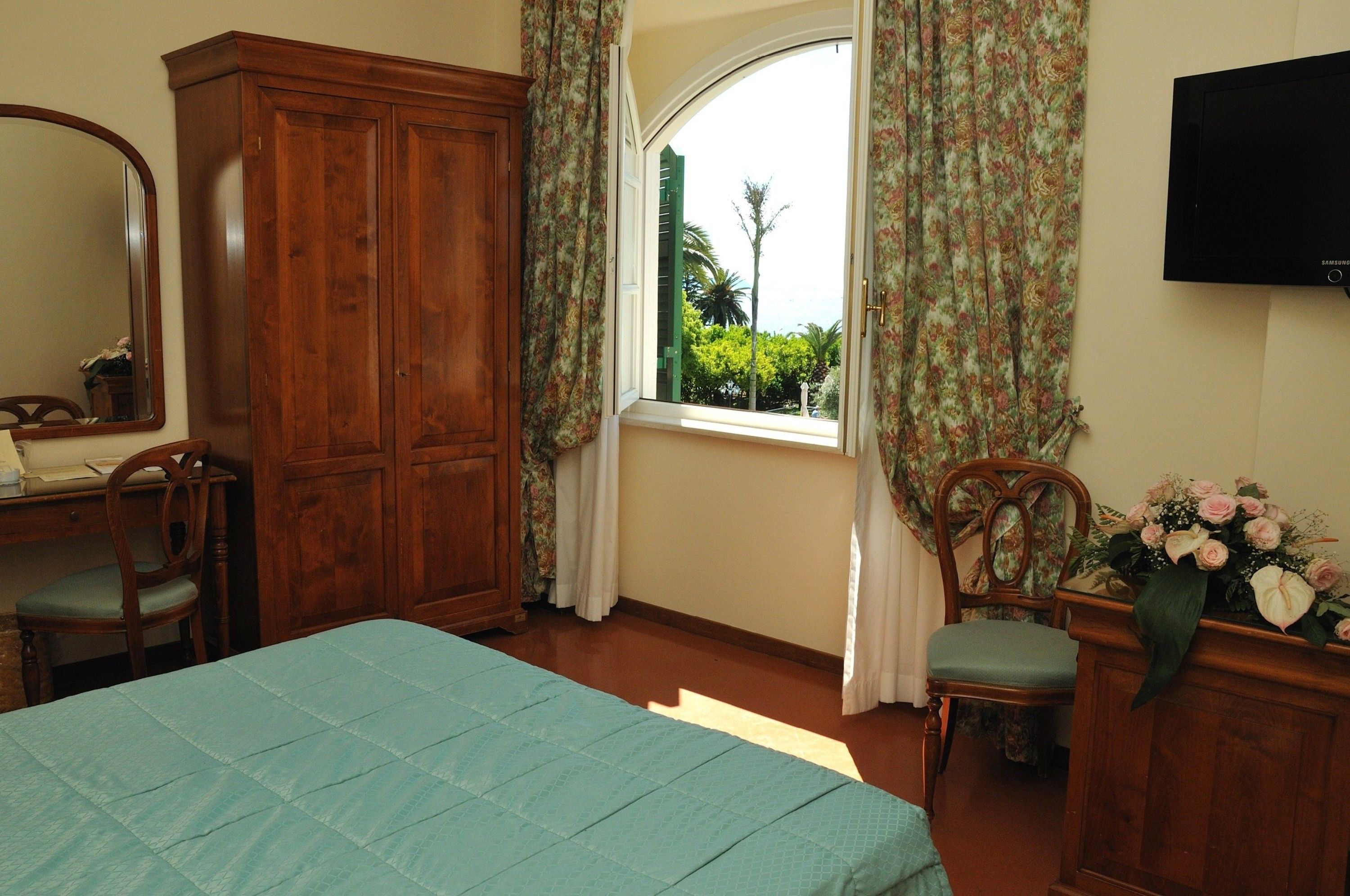 Une chambre à coucher comprenant un lit, une armoire en bois, un bureau avec une chaise, une fenêtre avec des rideaux et un téléviseur à écran plat fixé au mur.