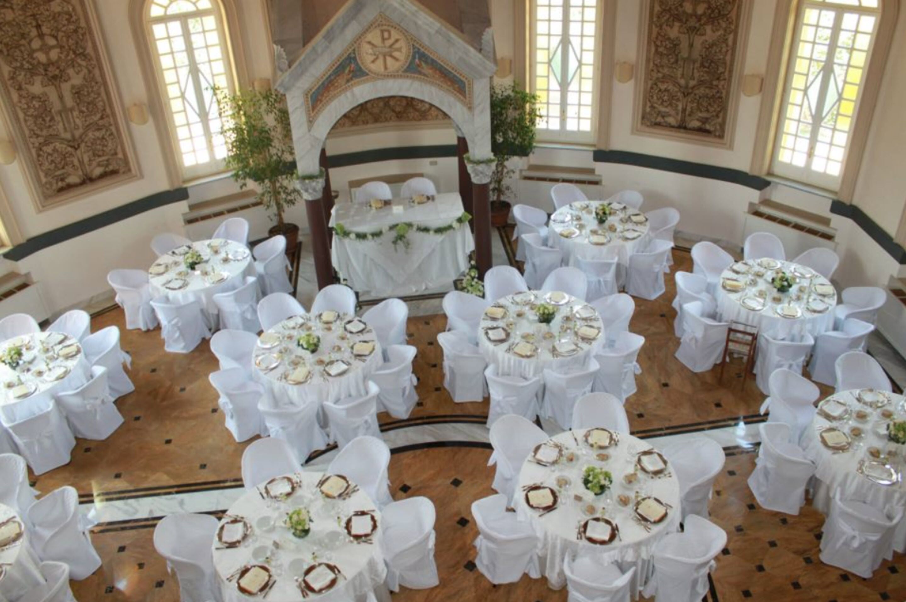 Un salón de banquetes con mesas redondas, sillas blancas y un escenario central con un dosel.