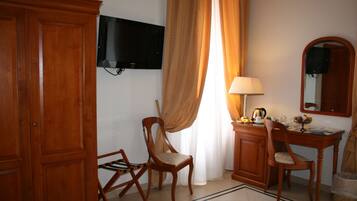 A hotel room with a wooden wardrobe, a desk with a lamp, a chair, a television mounted on the wall, and a window with curtains.