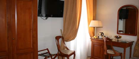 A hotel room with a wooden wardrobe, a desk with a lamp, a chair, a television mounted on the wall, and a window with curtains.