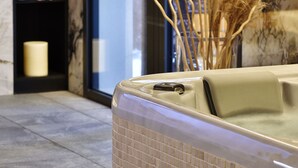 A spa area with a tiled floor, a decorative wall with a clock, a shelf with candles and towels, and a window with a view. - Hôtel Fesch & Spa (Ajaccio)