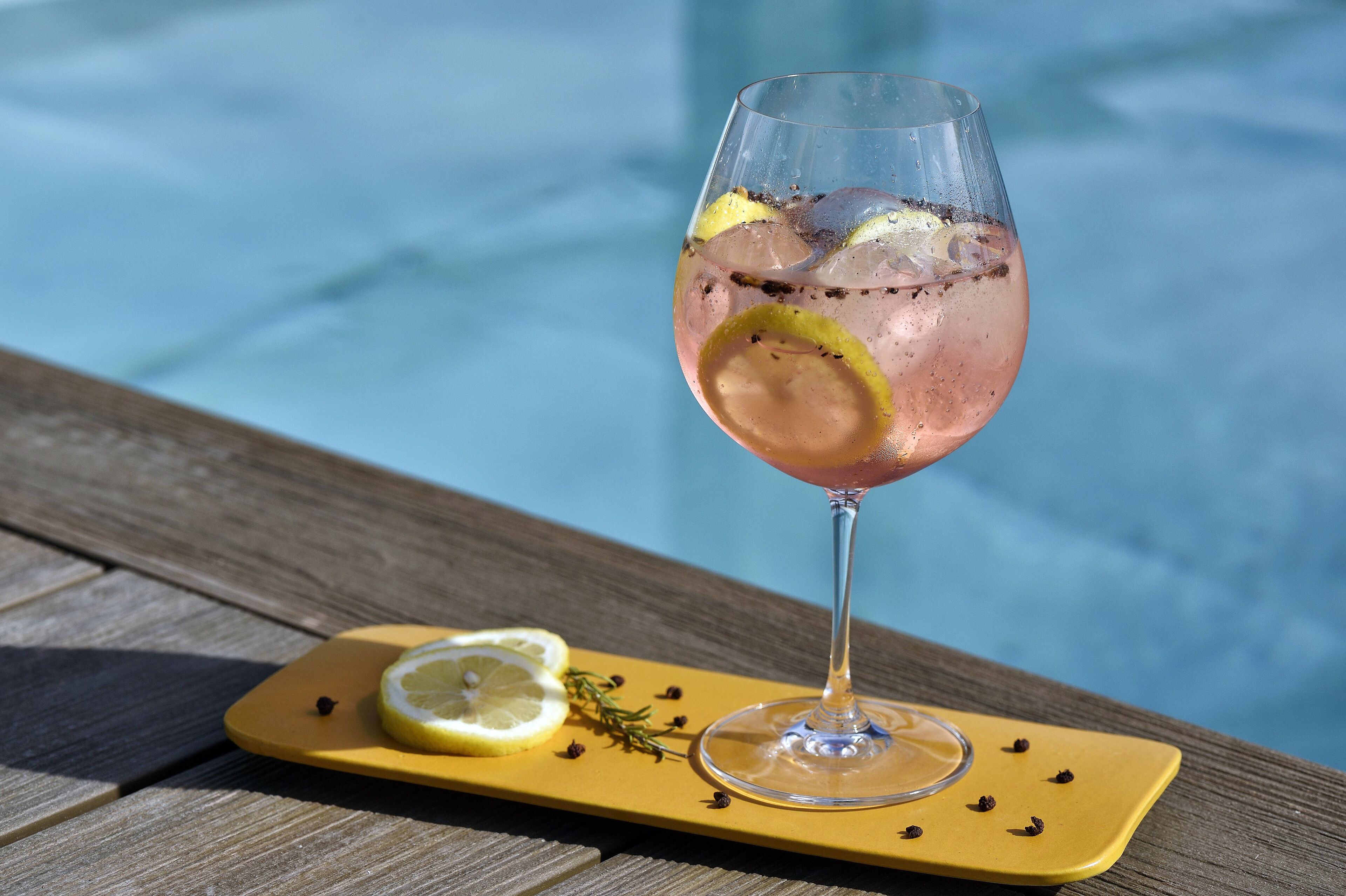 A glass of pink cocktail with lemon slices and ice, placed on a yellow tray with a slice of lemon and a sprig of rosemary beside it, by a pool.