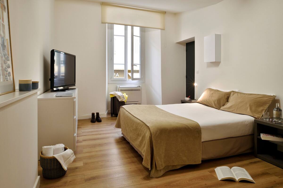 A hotel room with a bed, a TV, a window, and a bedside table.