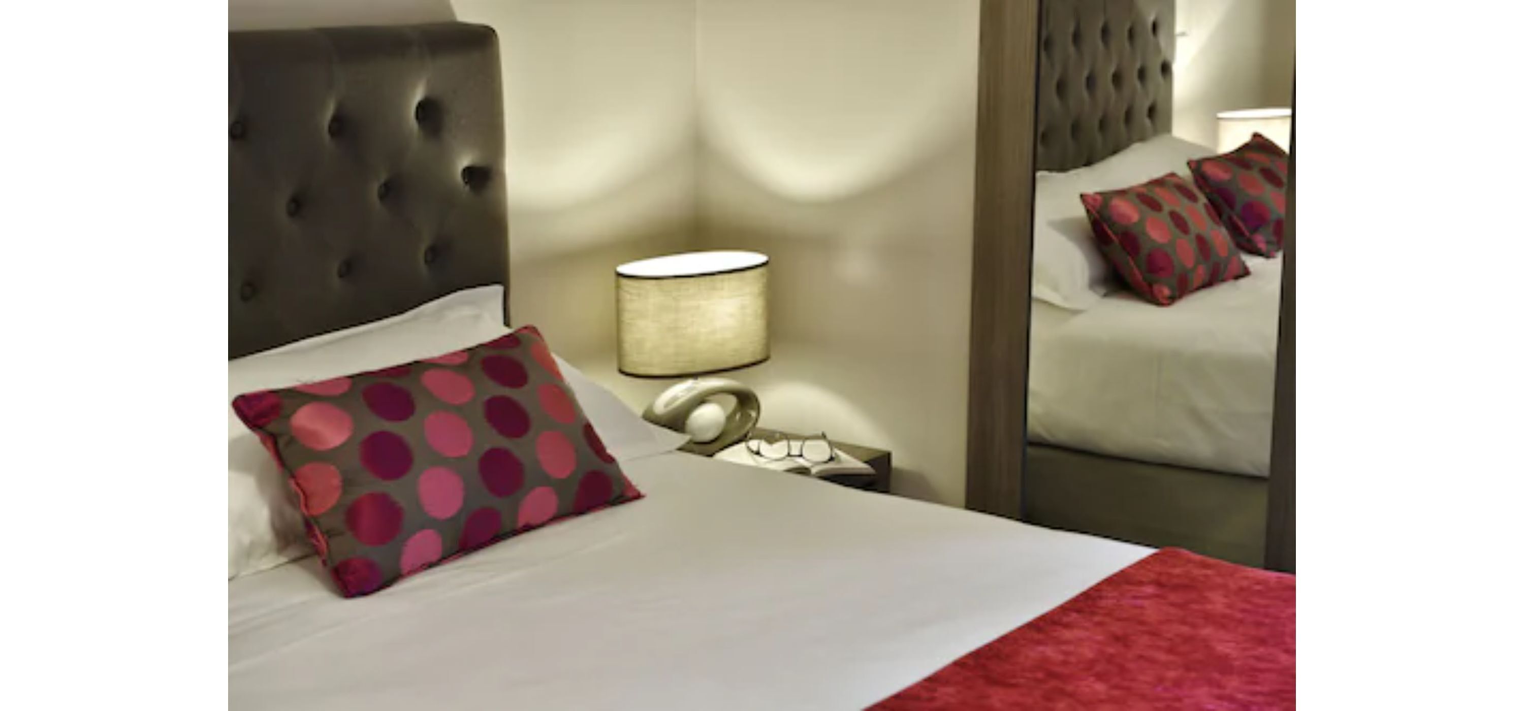 A hotel room with a tufted headboard, a bedside lamp, a polka dot pillow, and a mirror reflecting the bed and pillows.