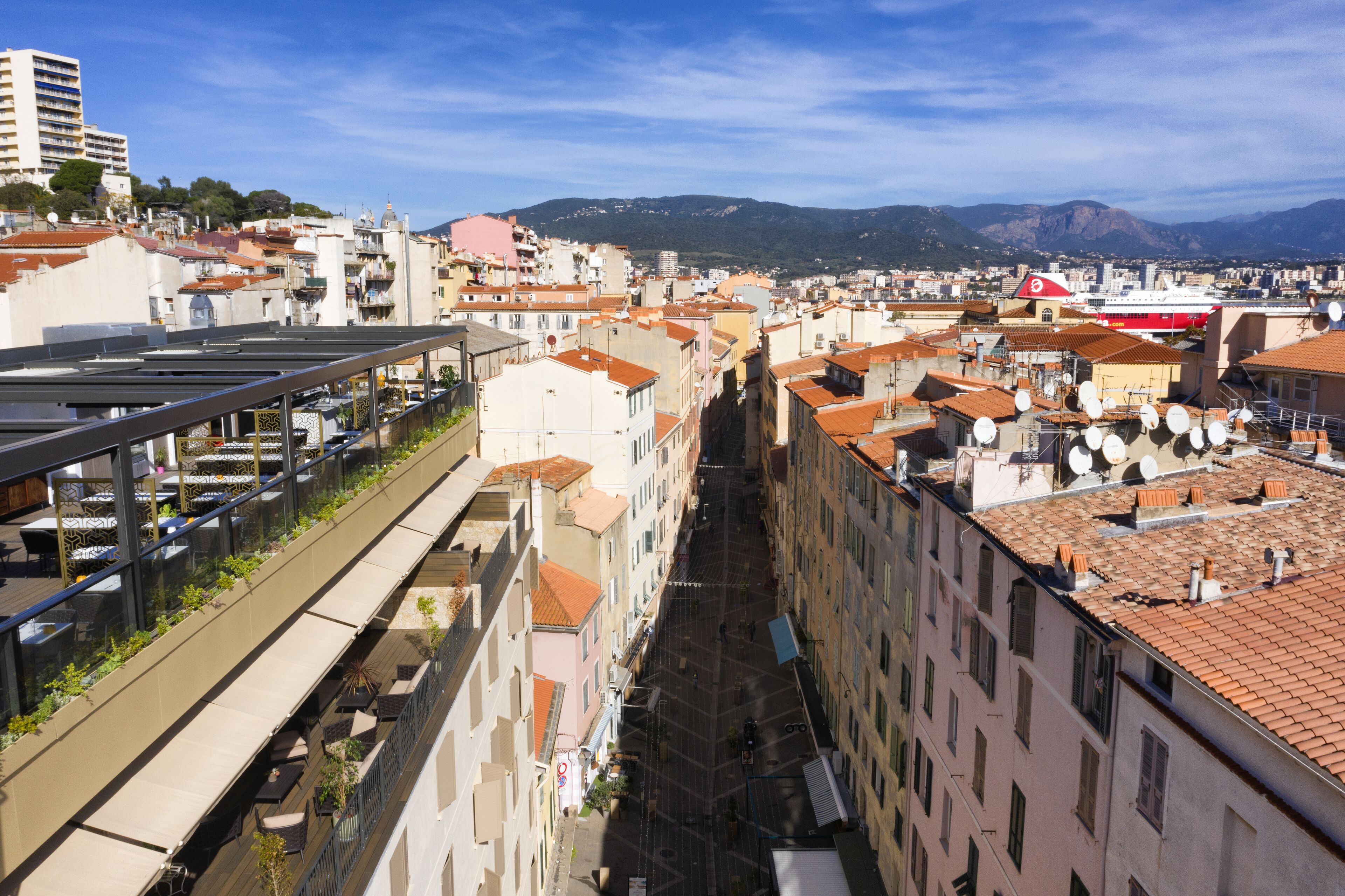 A narrow street flanked by buildings with terracotta roofs, a red and white boat docked on the water, and a clear blue sky.