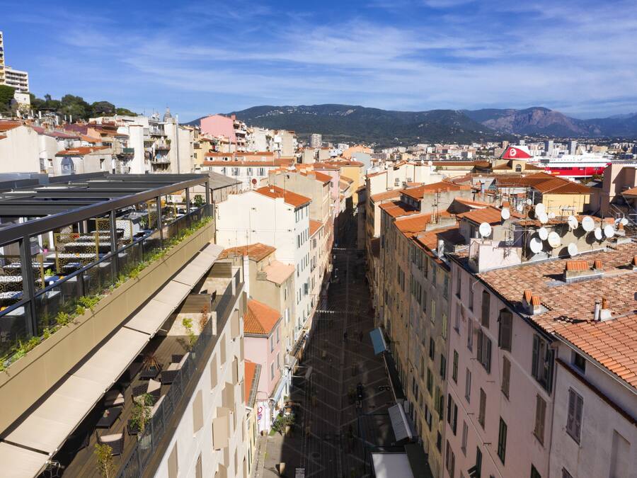 Une rue étroite bordée d’immeubles aux toits de tuiles rouges, un bateau rouge et blanc amarré sur l’eau, et un ciel bleu azur.