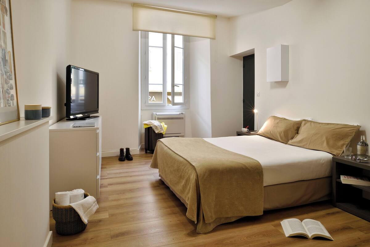 A bedroom with a bed, bedside table, TV, window, and a basket with towels.