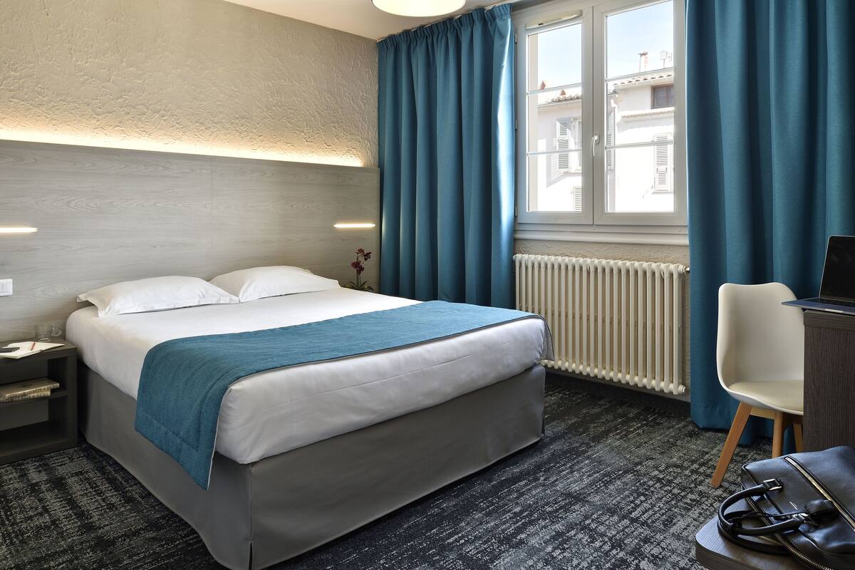 A hotel room with a bed, a desk with a chair, a radiator, and a window with curtains.