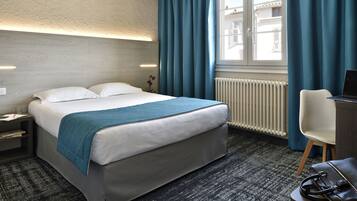 Une chambre d’hôtel avec un lit, un bureau et une chaise, un radiateur et une fenêtre avec des rideaux.