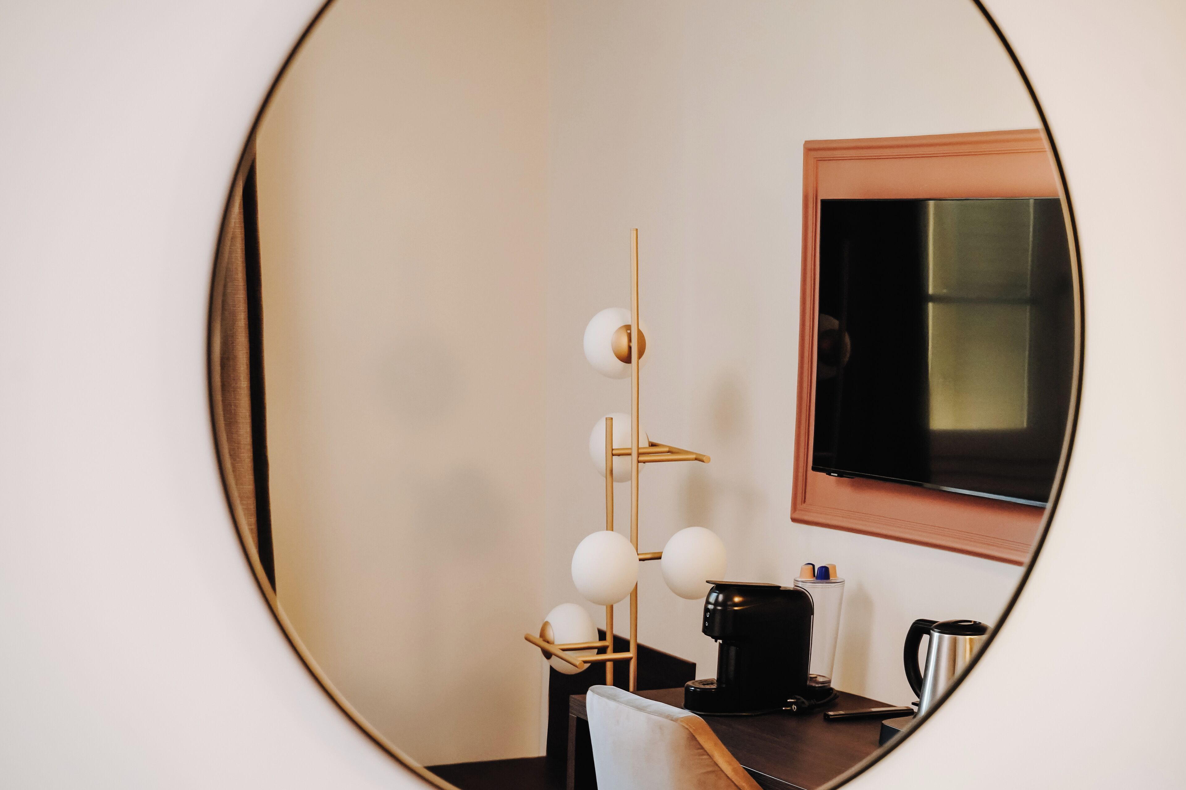 A room with a round mirror reflecting a modern lamp, a flat-screen TV, and a coffee maker.