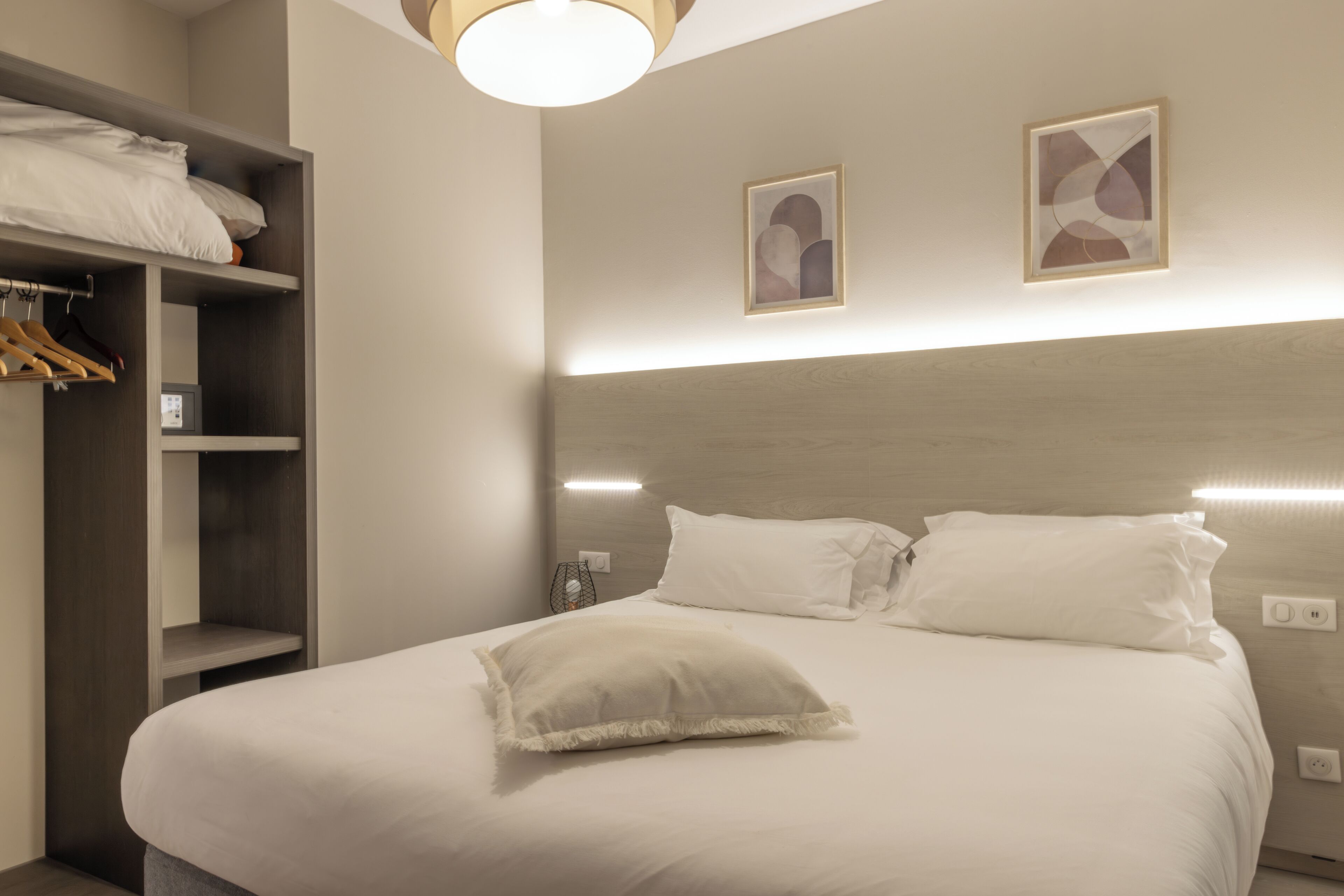 Een net opgemaakt bed met witte lakens en kussens, een houten hoofdbord en een wandlamp.