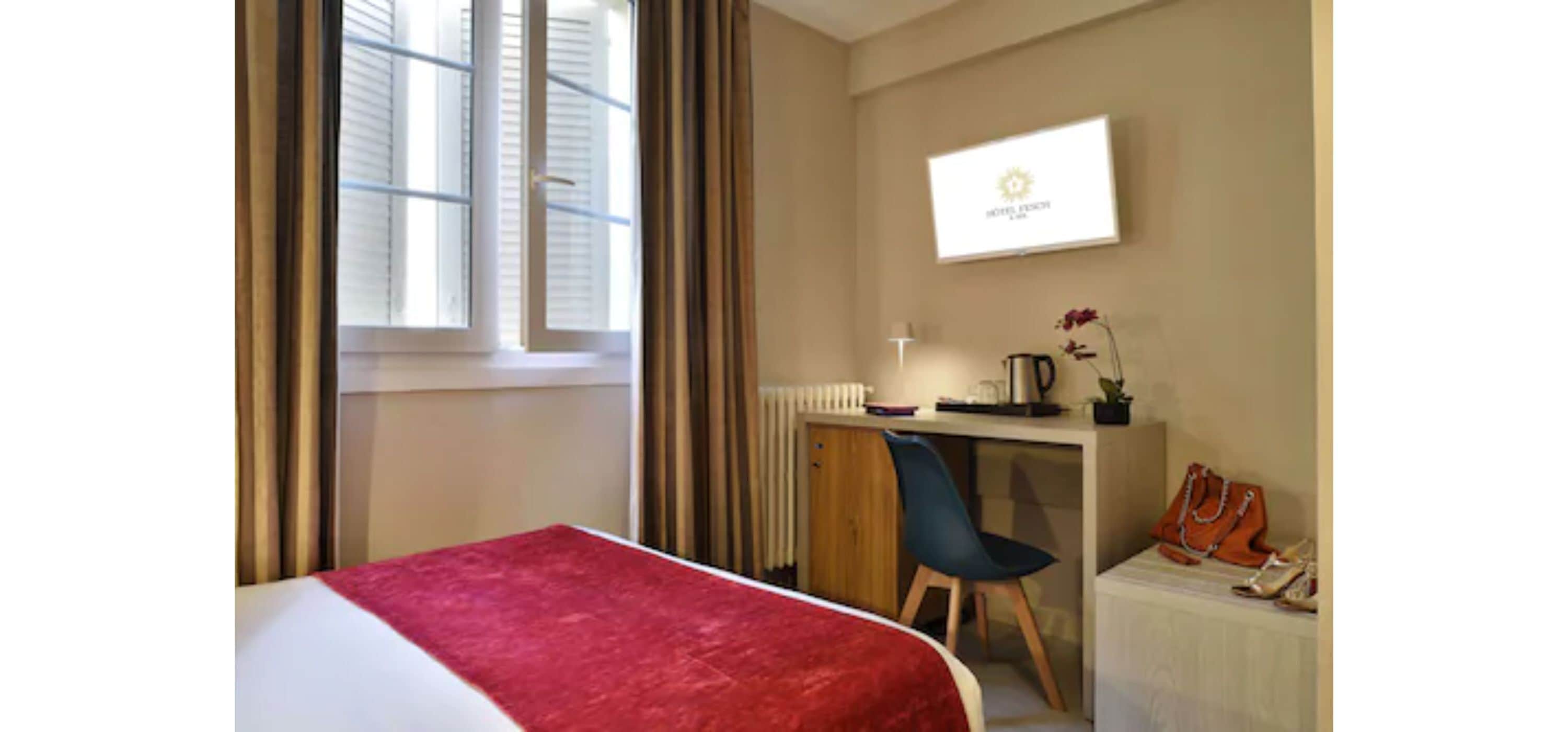 A hotel room with a bed, a desk, a chair, a radiator, and a TV on the wall.