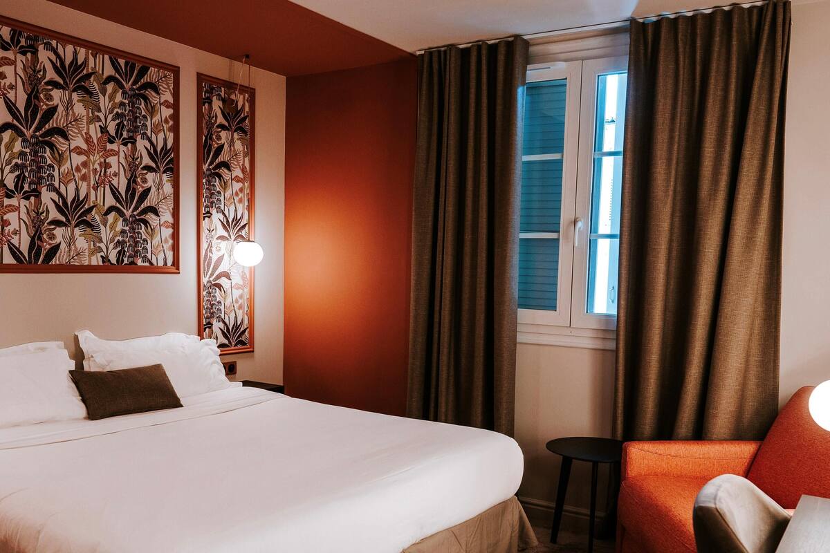 A hotel room with a large bed, a chair, a desk with a lamp, and a window with curtains.