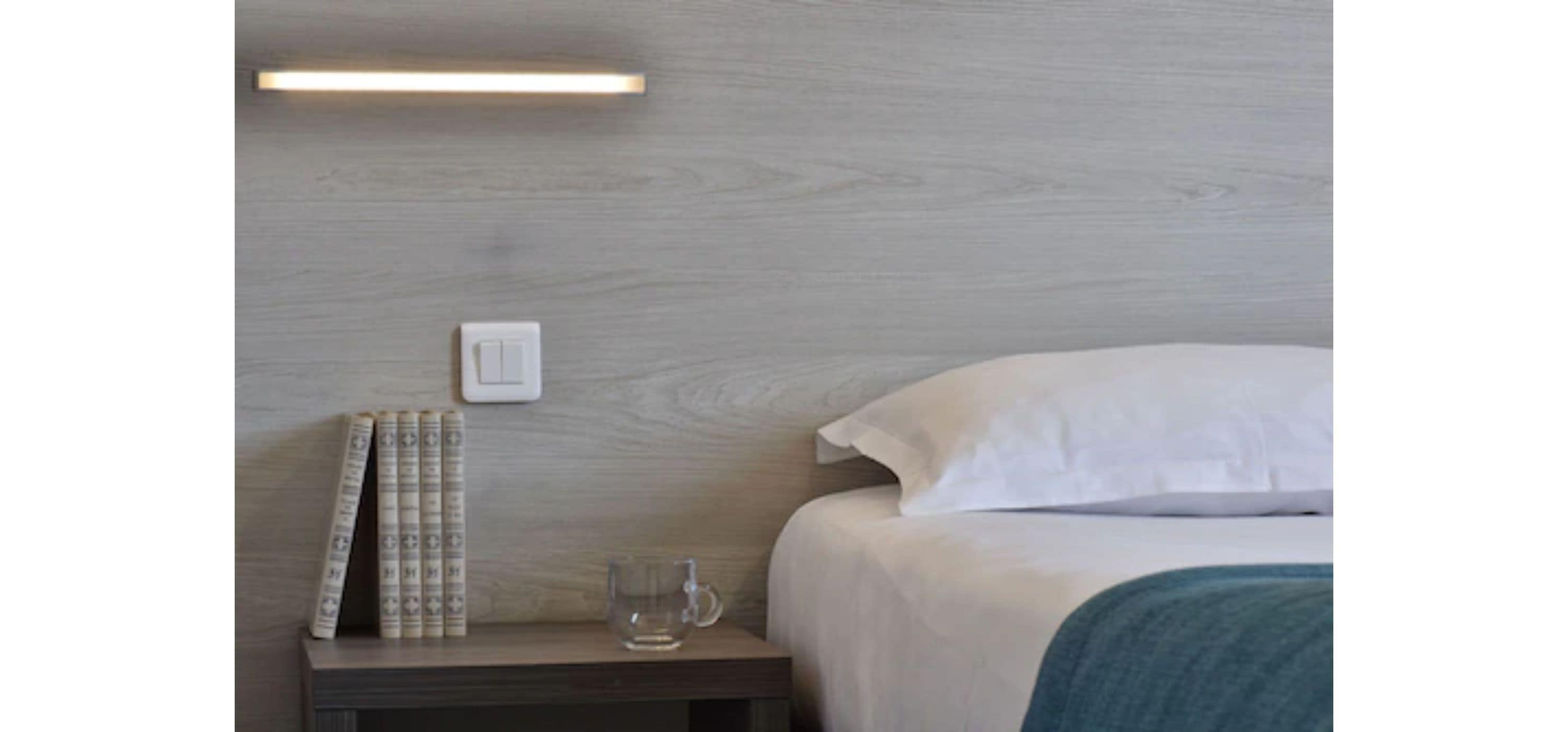 A neatly made bed with white sheets and a blue blanket, a bedside table with a glass mug, and a wall-mounted light fixture.