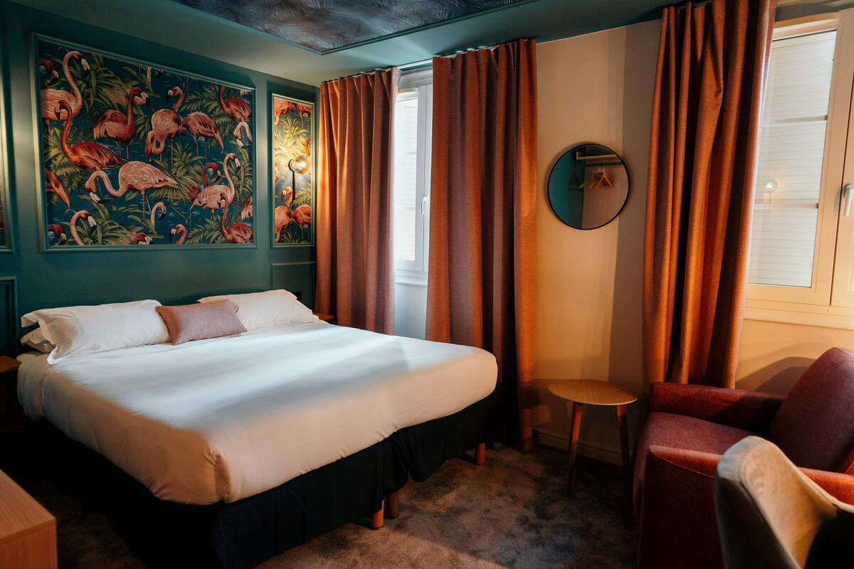 A hotel room with a large bed, a small table, a chair, and a painting of flamingos on the wall.