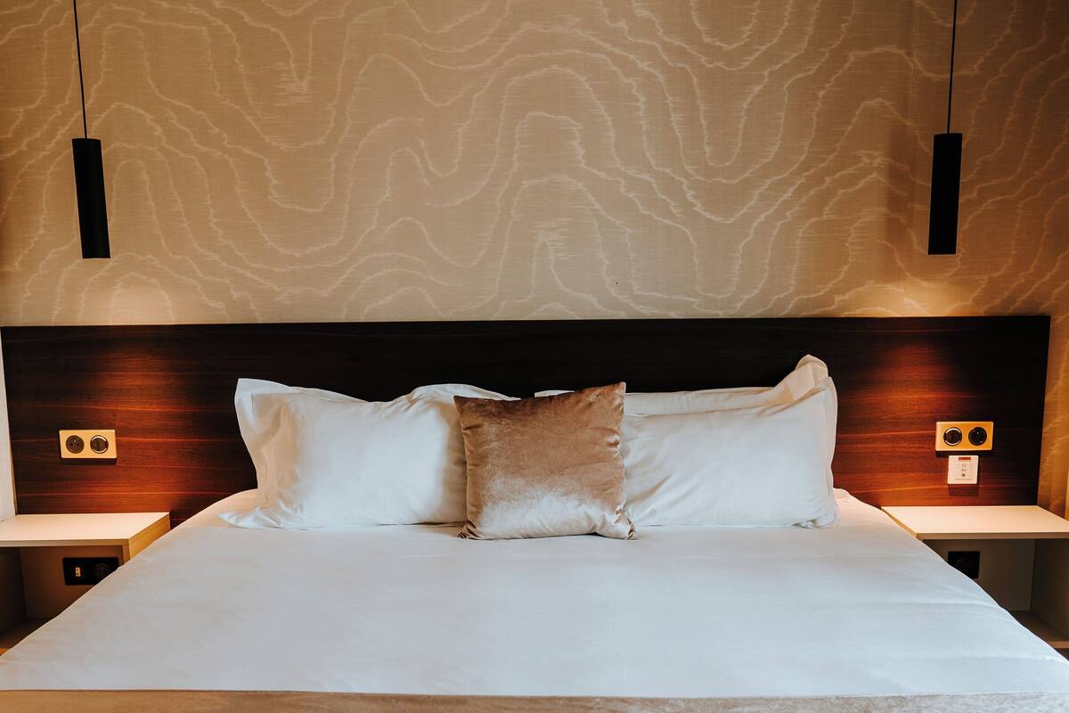 A neatly made bed with white linens and a brown throw blanket, flanked by two bedside tables with lamps, against a textured wall.